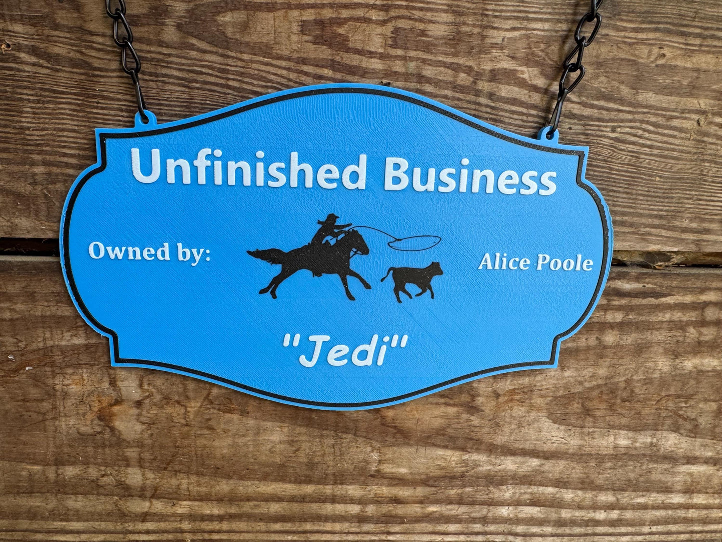 May include: Blue sign with white text that reads "Unfinished Business" Owned by: Alice Poole "Jedi". The sign has a black border and is hanging from a chain. The sign also features a silhouette of a person riding a horse and a calf.